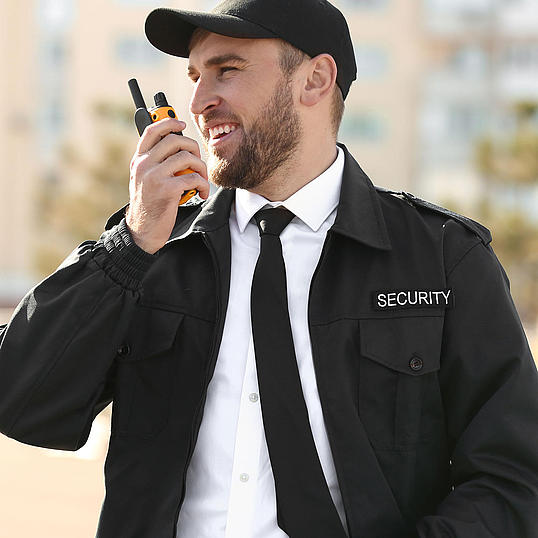 Security-Mitarbeiter Security-Mitarbeiter mit Cappy und Kravatte steht vor Gebäuden und spricht in ein Walkie-Talkie
