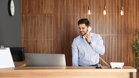 Junger dynamischer Mann in hellblauem Hemd steht hinter einem Empfangscounter und telefoniert.