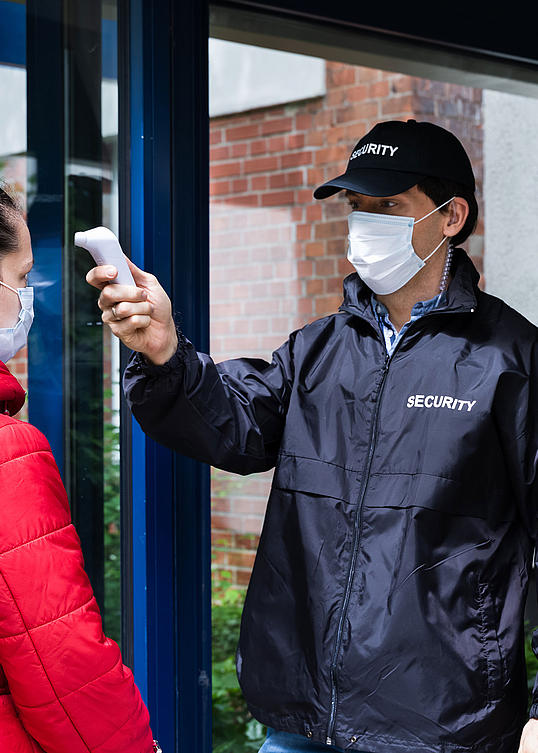 Security-Mann mit Cappy und Maske kontrolliert am Eingang die Körpertemperatur einer Frau mit Maske.