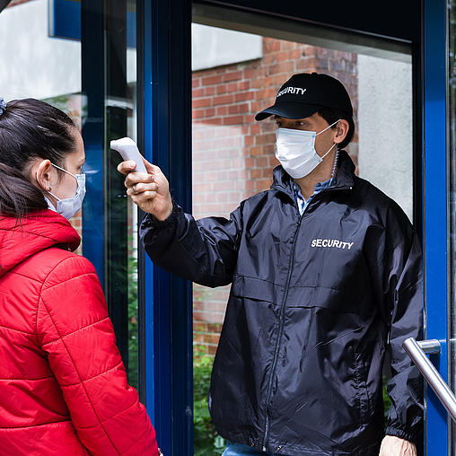 Security-Mann mit Cappy und Maske kontrolliert am Eingang die Körpertemperatur einer Frau mit Maske.