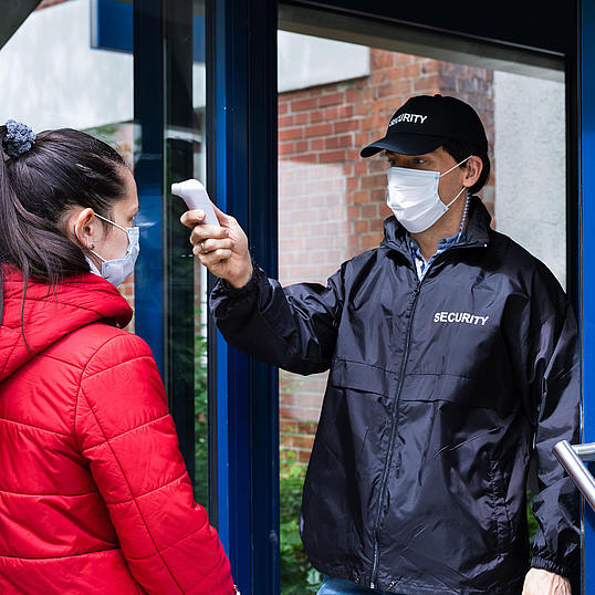 Security-Mann mit Cappy und Maske kontrolliert am Eingang die Körpertemperatur einer Frau mit Maske.
