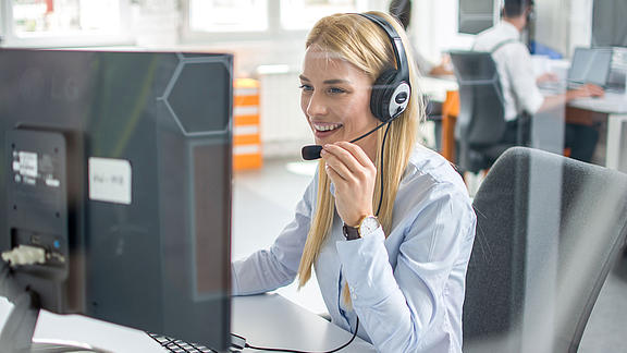 Blonde, langhaarige Frau sitzt mit einem Headset vor einem Bildschirm und beantwortet Telefonate