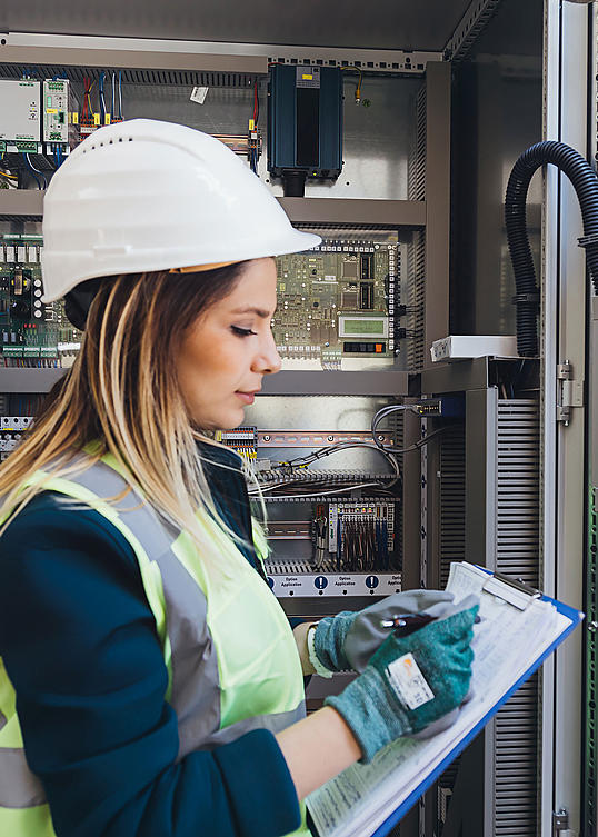 Frau mit Handschuhen, Warnweste und Helm kontrolliert Elektrik und macht Notizen auf einem Klemmbrett.