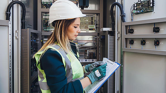 Frau mit Handschuhen, Warnweste und Helm kontrolliert Elektrik und macht Notizen auf einem Klemmbrett.