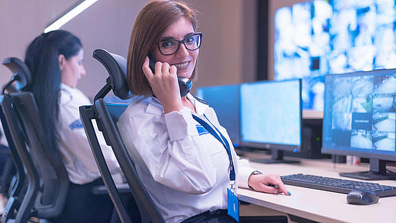 Junge Frau mit Blouse und Brille sitzt an einem Schreibtisch in einer Überwachungszentrale und telefoniert.